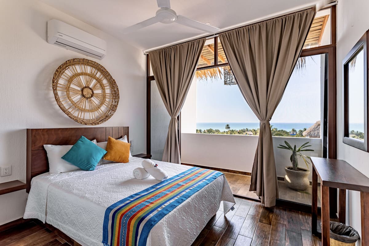 A bedroom with a large bed, a ceiling fan, a view of the ocean, and a potted plant.