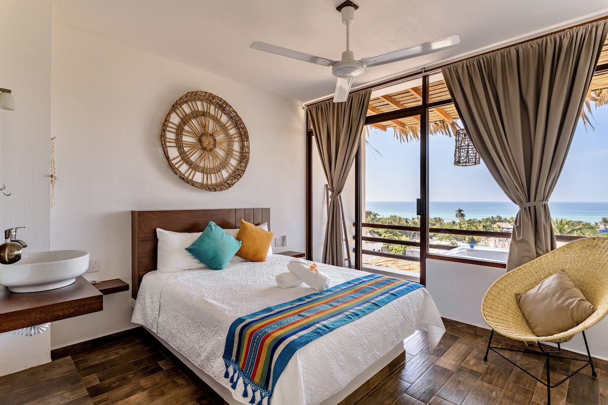 A bedroom with a bed, a ceiling fan, a large window with a view of the ocean, a wicker chair, and a wall-mounted decorative item.