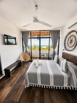 Un dormitorio con cama, televisor, ventilador de techo, vista al océano y balcón con una silla.