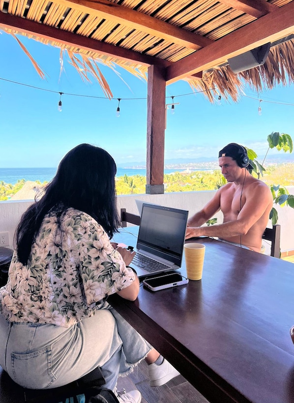 A person using a laptop at a wooden table with a view of the ocean. - PUNTA KAI  (Puerto Escondido)