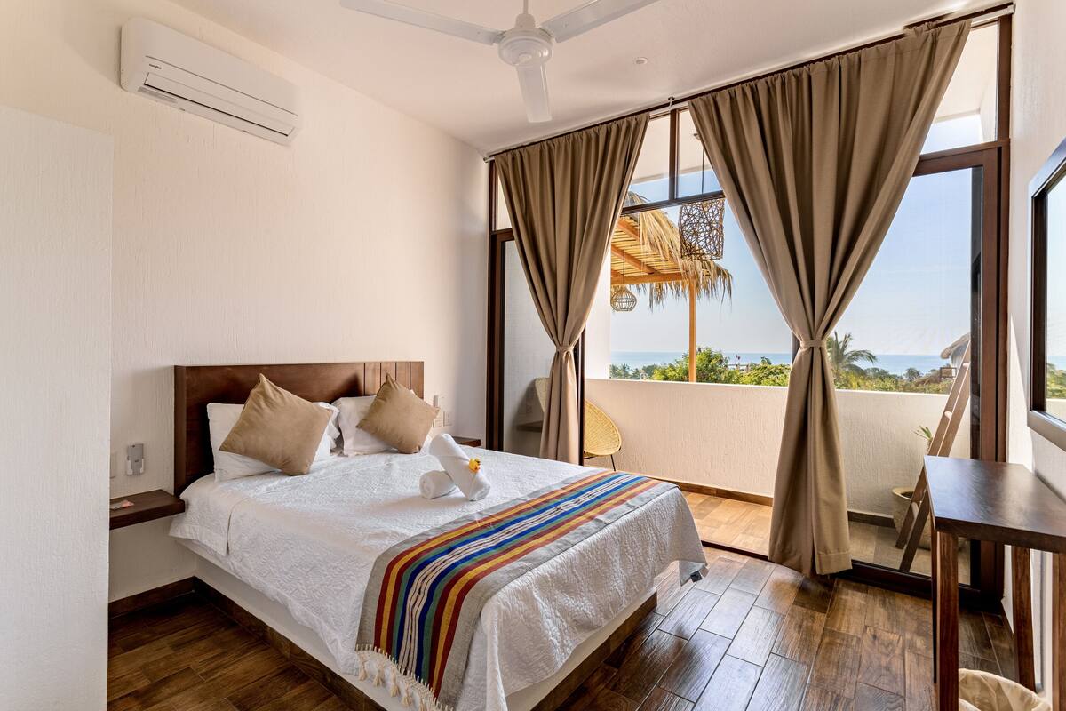 A hotel room with a bed, a ceiling fan, a window with curtains, and a view of the outdoors.