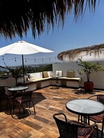 Un patio en la azotea con techo de paja, zona de estar, luces colgantes y macetas con plantas.