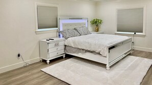 Un dormitorio con cama, mesitas de noche, una mesita con un florero y dos ventanas con persianas.