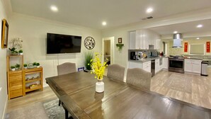 Una sala moderna con una mesa de comedor de madera, un televisor de pantalla plana montado en la pared y una zona de cocina con armarios blancos y electrodomésticos de acero inoxidable.