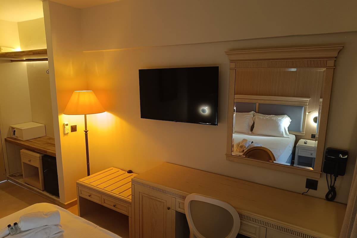 Una habitación de hotel con cama, escritorio, silla, televisor, lámpara, espejo y mesita de noche.