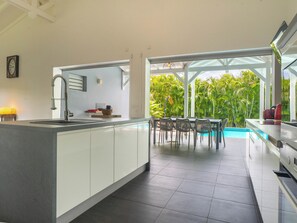 Une cuisine moderne dotée d’un grand îlot central, d’un coin repas avec des chaises, et offrant une vue sur une piscine et une végétation luxuriante.