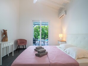A bedroom with a bed, a desk, a chair, a nightstand, and a view of a pool and greenery outside.