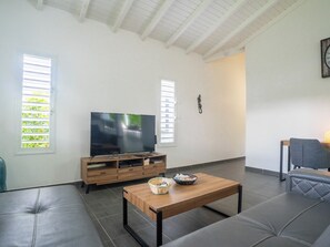 A living room with a flat-screen TV on a wooden stand, a coffee table, a sofa, and two windows with blinds.