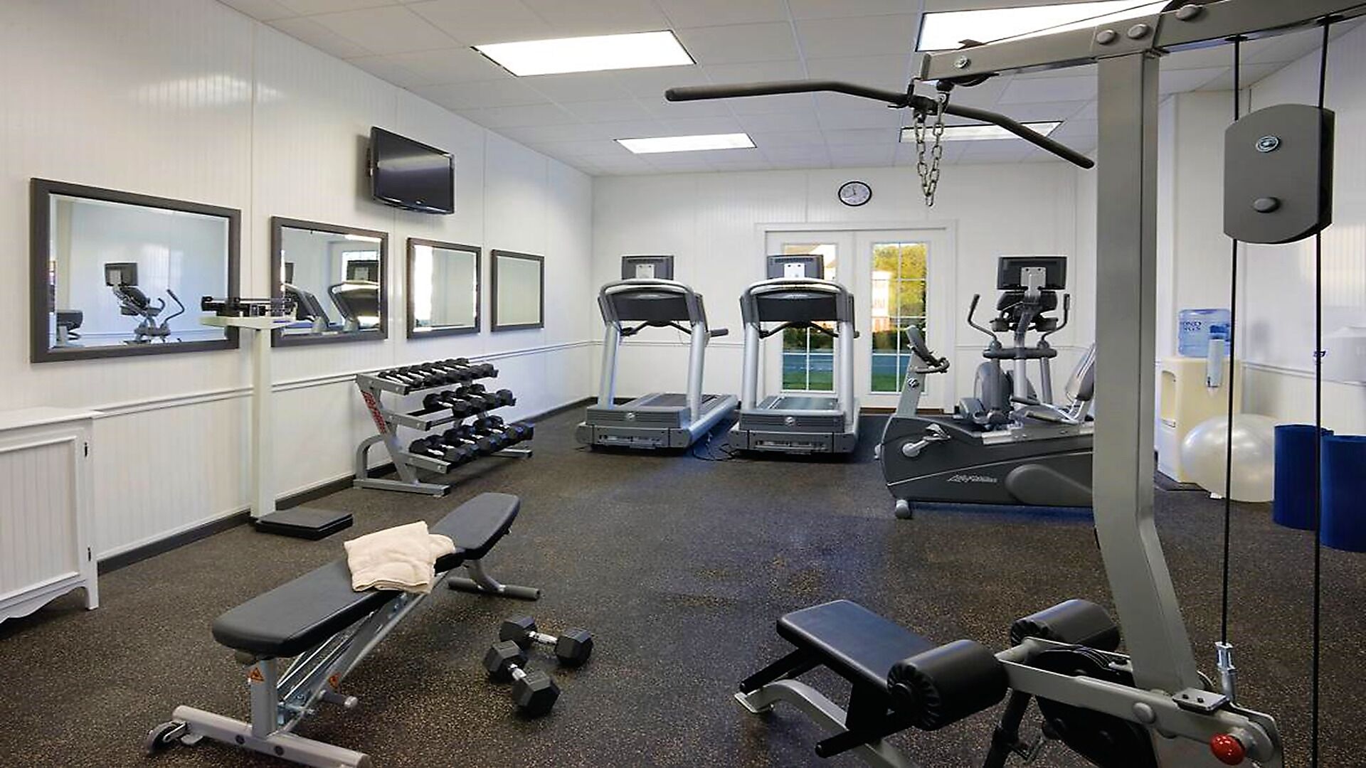 Une salle de sport équipée de tapis de course, de vélos elliptiques, d’haltères et de bancs.