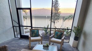 Een balkon met rieten meubilair, een glazen tafel en plantenbakken.
