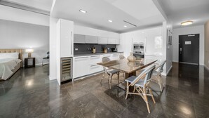 A modern kitchen with white cabinets, a dining area with a wooden table and wicker chairs, and a bed with a headboard in the adjacent room.