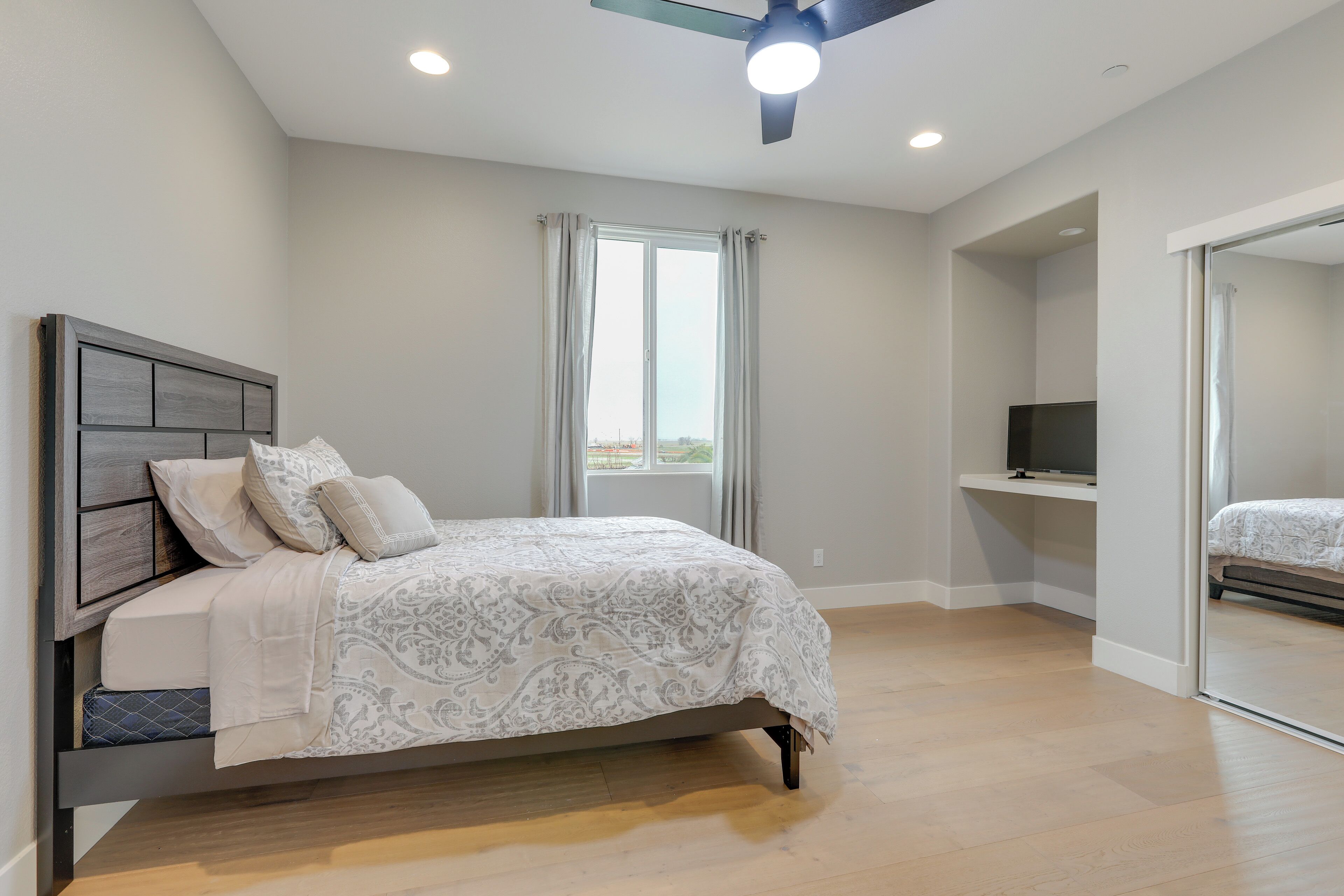 Um quarto com cama, ventilador de teto, janela com cortinas e TV em uma estante.