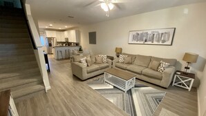 A modern living room with a sofa set, coffee table, and patterned rugs. A kitchen and dining area are visible in the background. - Set Sail 7 - Spacious Townhome 3 Bed/2.5 bath/sleeps7 - 1 min walk to the beach! (Mexico beach)