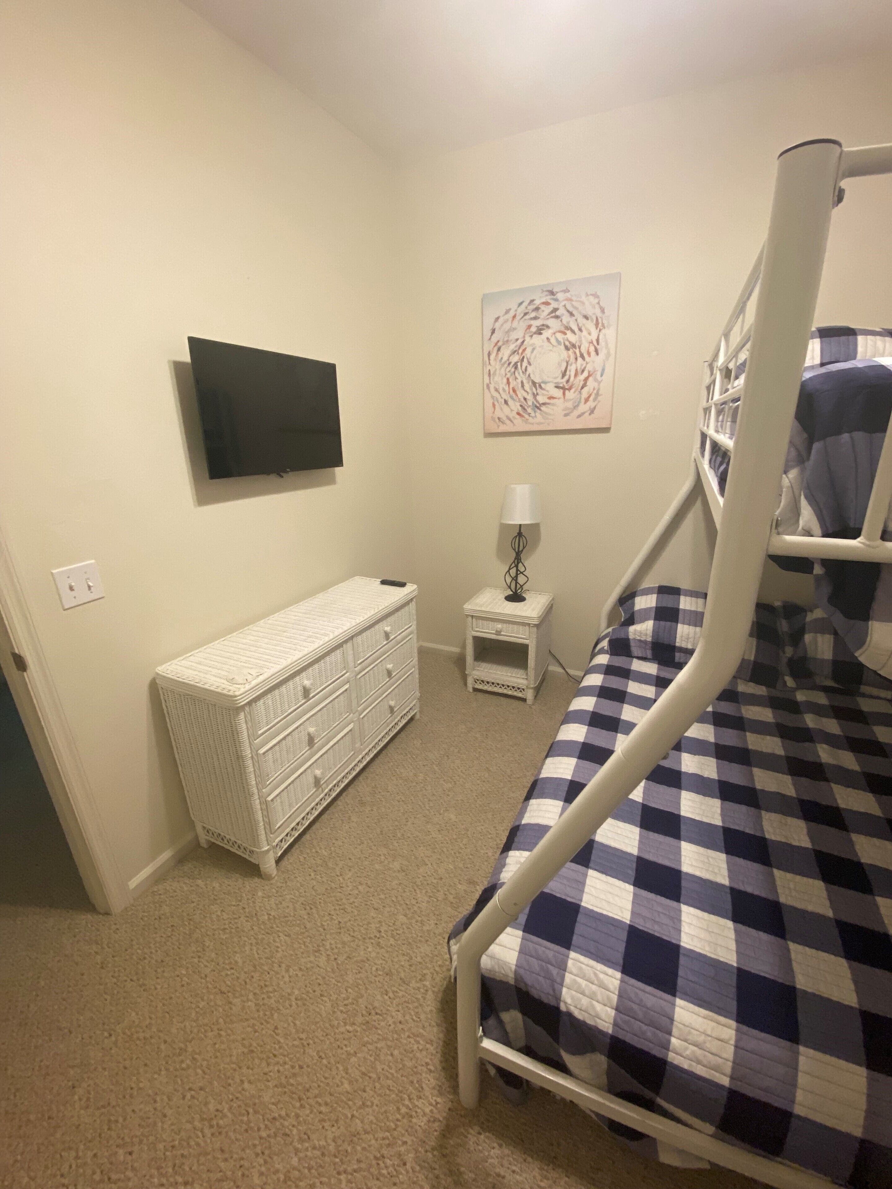 Een slaapkamer met een stapelbed, een witte dressoir, een klein nachtkastje met een lamp en een aan de muur bevestigde televisie.