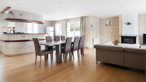 Une cuisine moderne dotée d’un îlot central aux lignes courbes, un coin repas avec une grande table et des chaises, et un espace de vie comprenant un canapé et une cheminée.