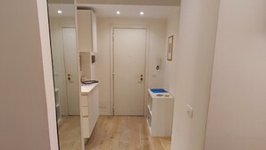 Un pasillo estrecho con suelo de madera, una puerta blanca al fondo y una pequeña zona de kitchenette con encimera azul.