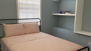 Ein Schlafzimmer mit einem Bett, einem Fenster mit Jalousien, einem Ablagefach mit Handtüchern und einem an der Wand befestigten Regal.