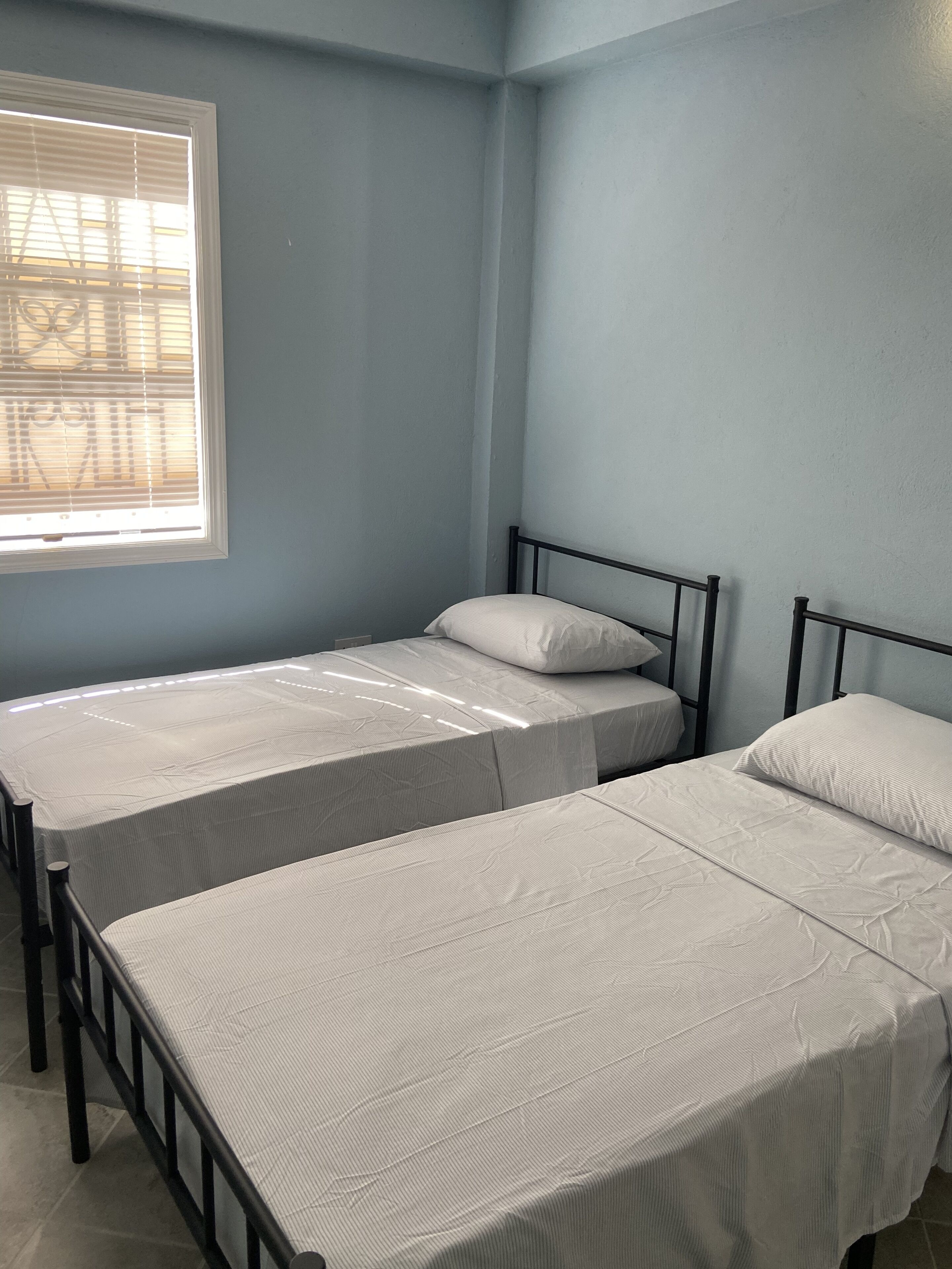 Een kamer met twee eenpersoonsbedden, een raam met zonwering en lichtblauwe wanden.