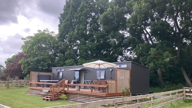 Eine moderne, kompakte Blockhütte mit einer Terrasse und Sitzgelegenheit im Freien, umgeben von einem Holzzaun und üppiger Bepflanzung.