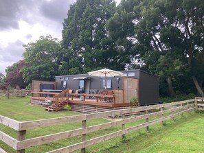 A modern, compact cabin with a deck and outdoor seating area, surrounded by a wooden fence and lush greenery. - Country tiny house with spa (Arapuni)