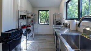 A compact kitchen with metal shelving, a stainless steel sink, and a window with a forest view. - The Glo Haus @ Four Fillies Lodge (Peterstown)