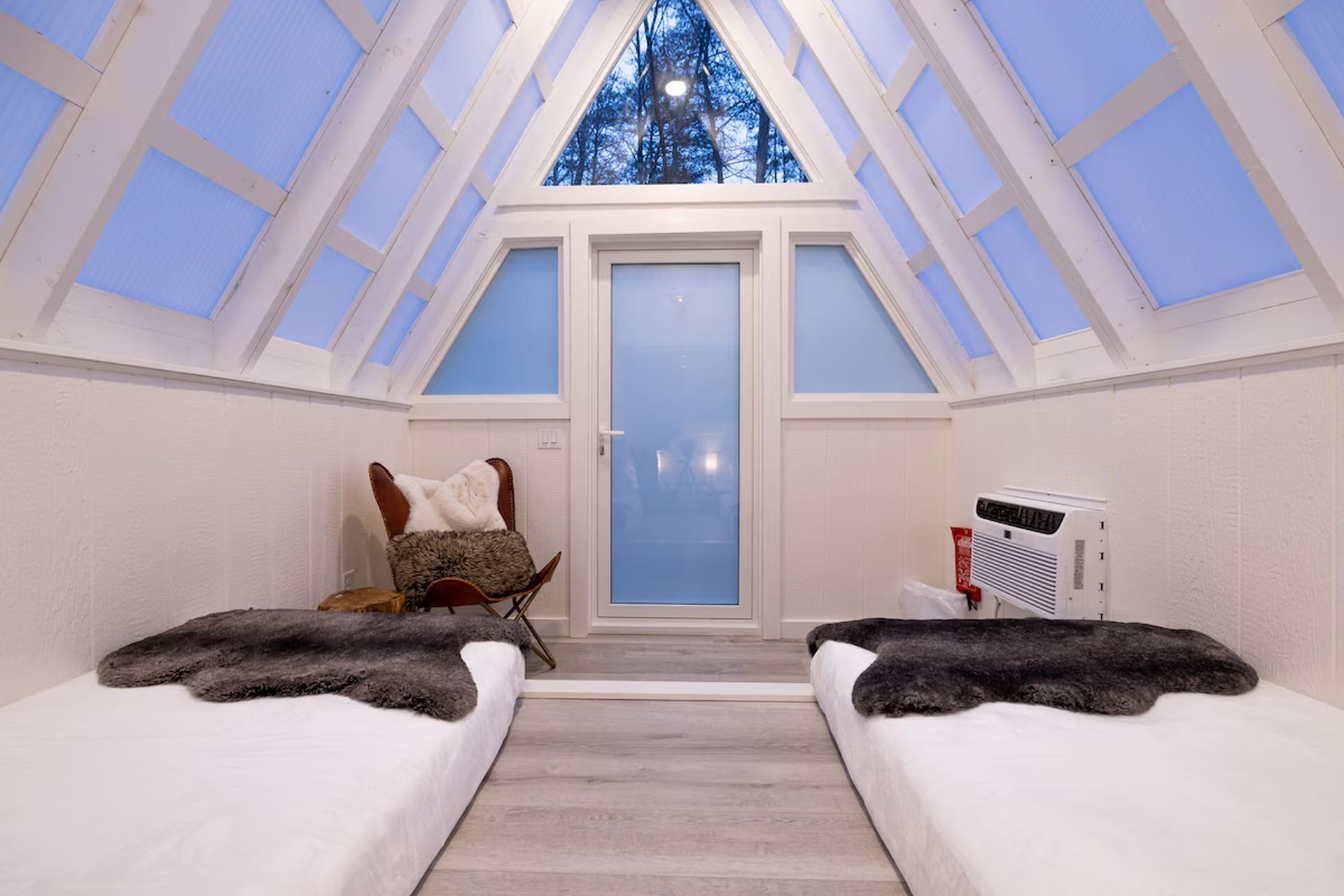A room with a sloped ceiling, a door, two beds with fur throws, a chair with a fur throw, and a heater.