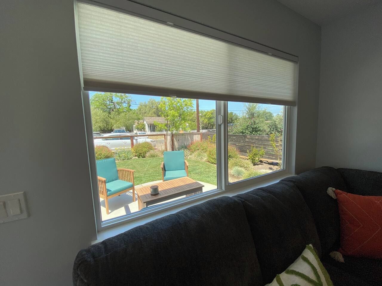 Una habitación con un ventanal grande, un sofá de color oscuro y vistas a un patio trasero con terraza de madera, sillas y follaje.