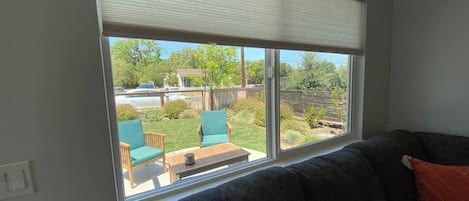 A room with a large window, a dark-colored sofa, and a view of a backyard with a wooden deck, chairs, and greenery.