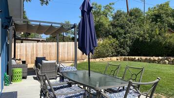 Un patio al aire libre con mesa y sillas de vidrio, una zona de estar cubierta y una cerca de madera al fondo.