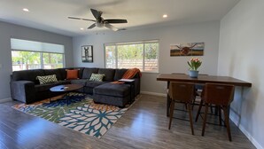 A modern living room with a sectional sofa, a dining table with chairs, a ceiling fan, and large windows.