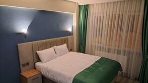 Ein ordentlich bezogenes Bett mit einer grünen Tagesdecke, ein Nachttisch mit Schubladen und eine Wandleuchte.