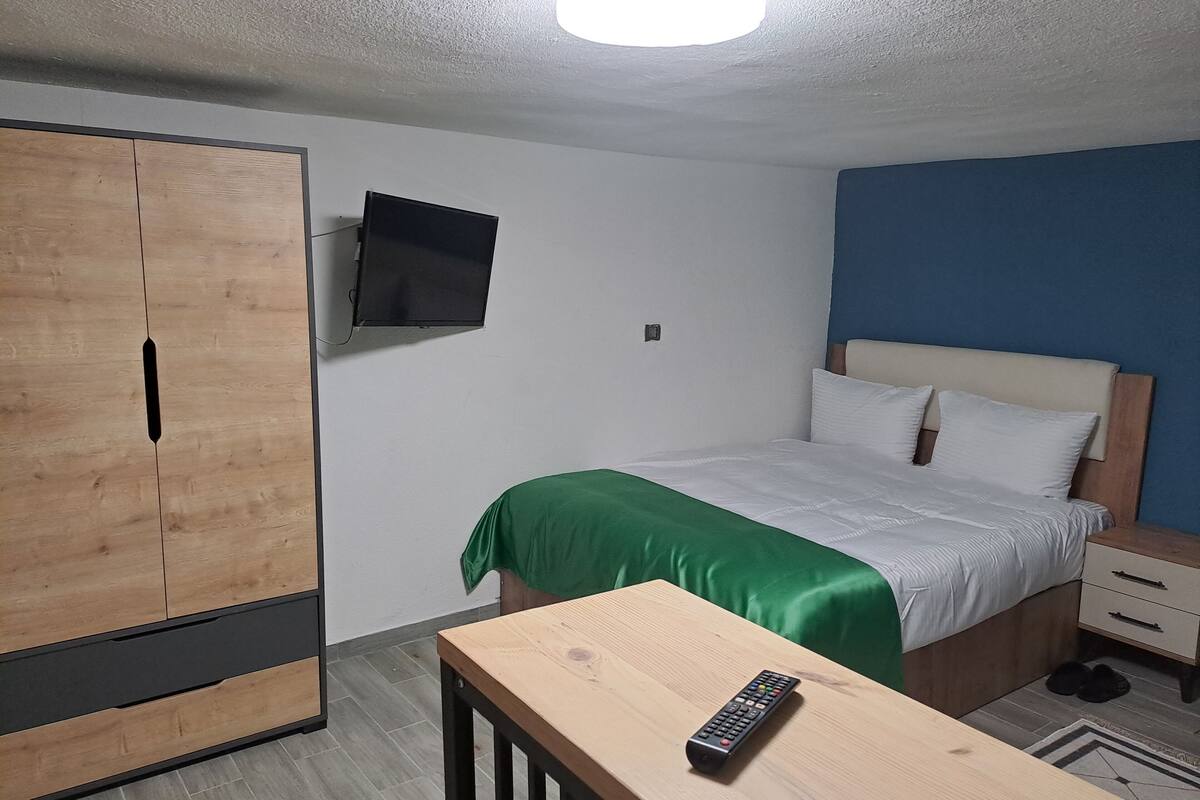 A bedroom with a wooden wardrobe, a bed with a green bedspread, a TV mounted on the wall, and a wooden nightstand with a drawer.