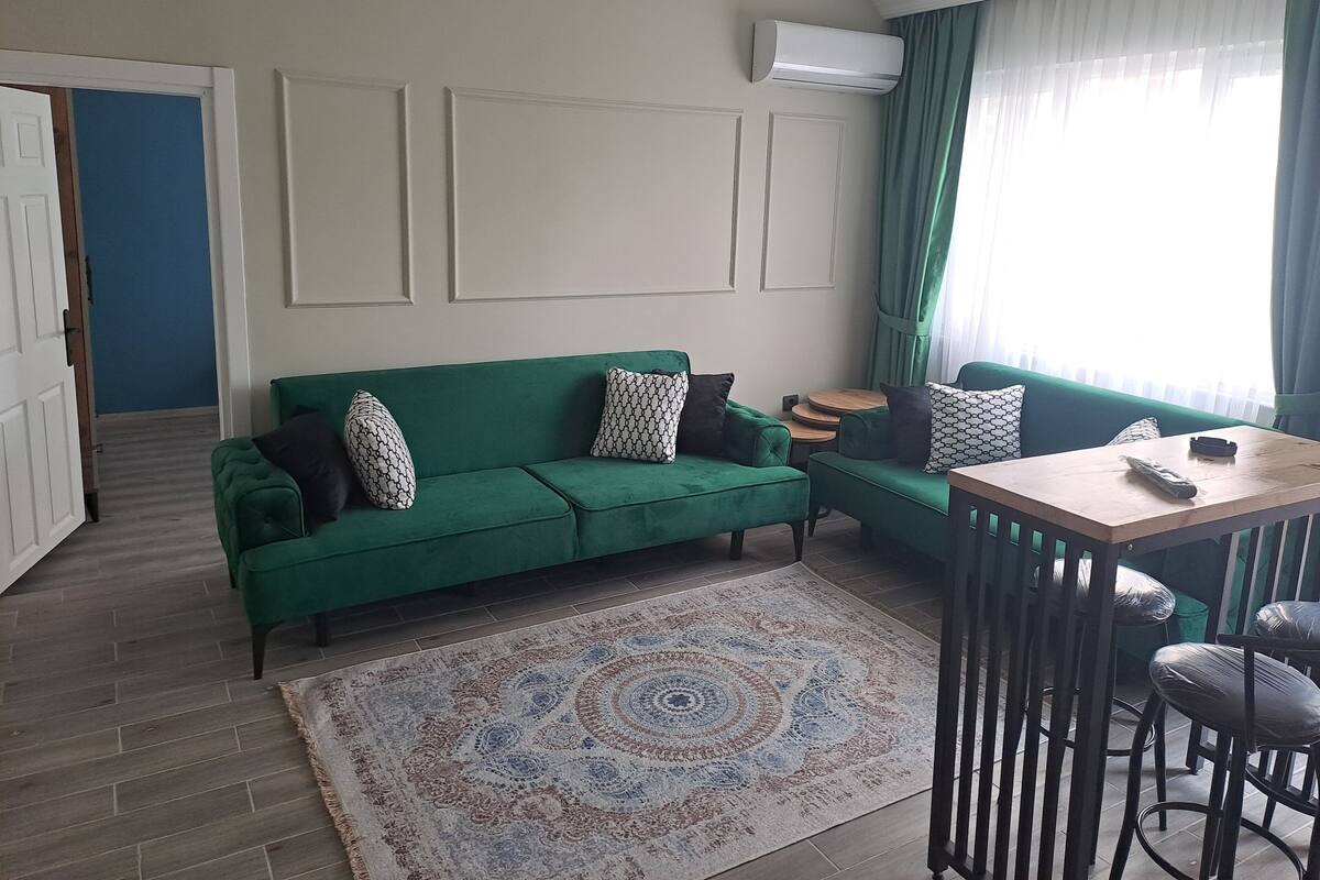 A green sofa with patterned cushions, a small wooden table, and a bar stool.