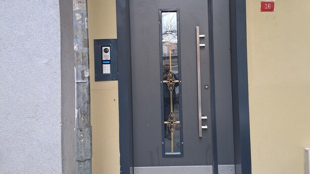 Eine dunkelgraue Tür mit vertikalem Griff und einem goldenen Zierteil, ein kleines elektronisches Gerät an der linken Wand und ein rotes Schild mit der Nummer 16 an der rechten Wand.