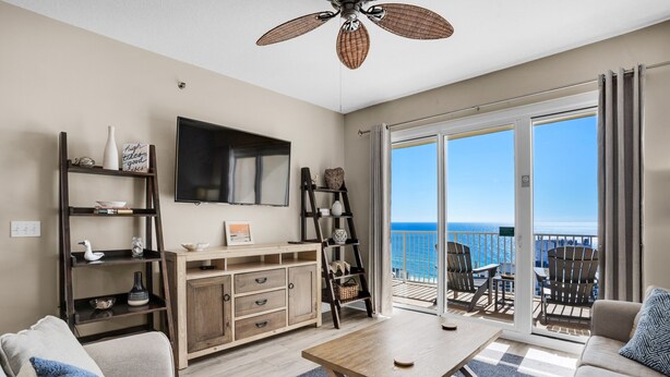 A living room with a large TV, wooden furniture, a balcony with chairs, and a view of the ocean.