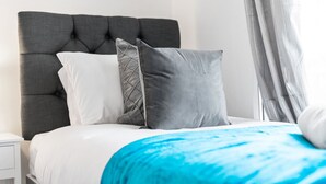 Ein ordentlich bezogenes Bett mit grauem Kopfteil, weißen und grauen Kissen und einer blauen Decke.