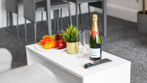 Ein weißer Tisch mit einer Champagnerflasche, zwei Gläsern, einer Fernbedienung und einer Obstschale mit einer Topfpflanze.