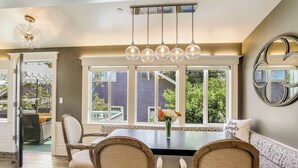 A dining area with a large table, four chairs, a chandelier, and large windows with a view of the outside.