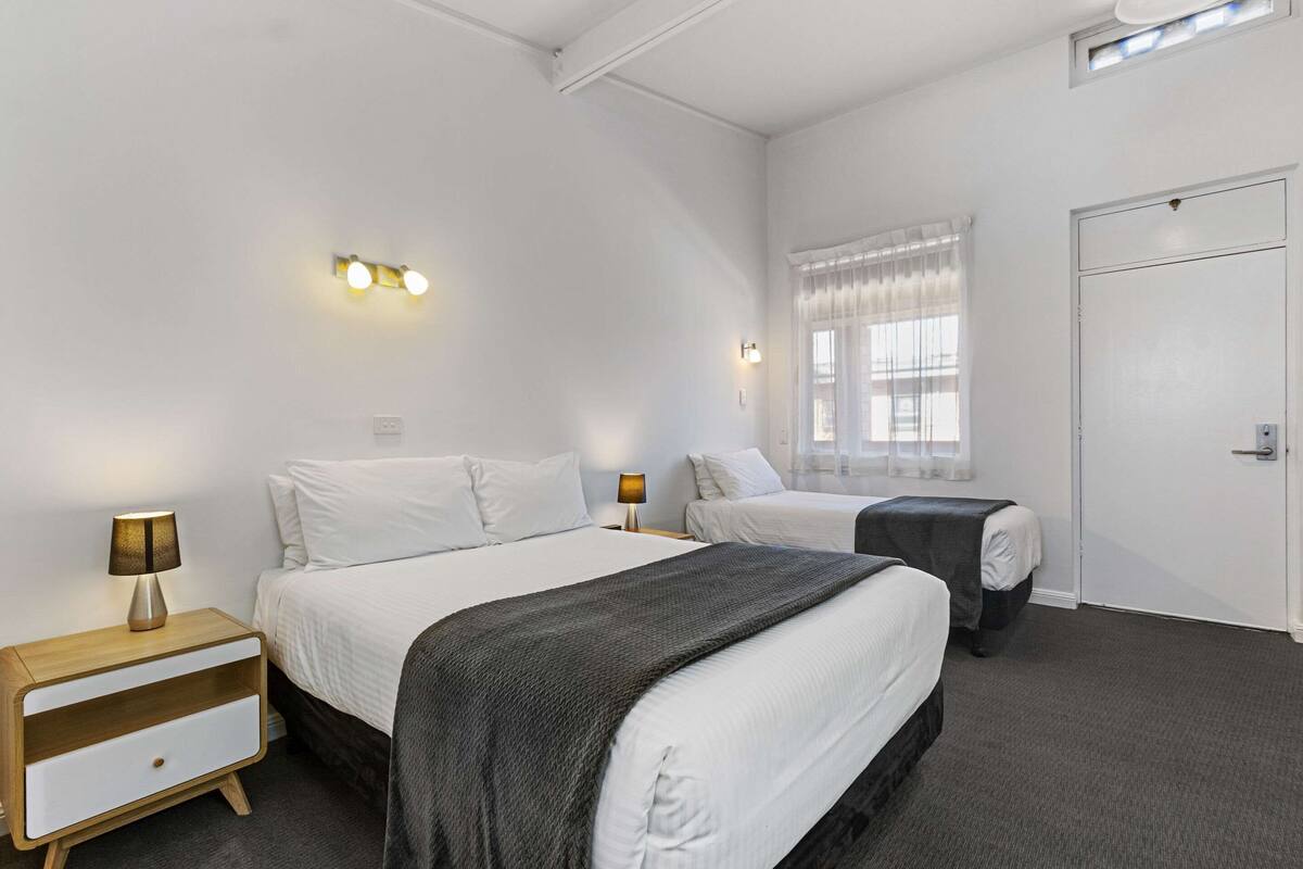 A hotel room with two beds, a nightstand with a lamp, a window with sheer curtains, and a door.