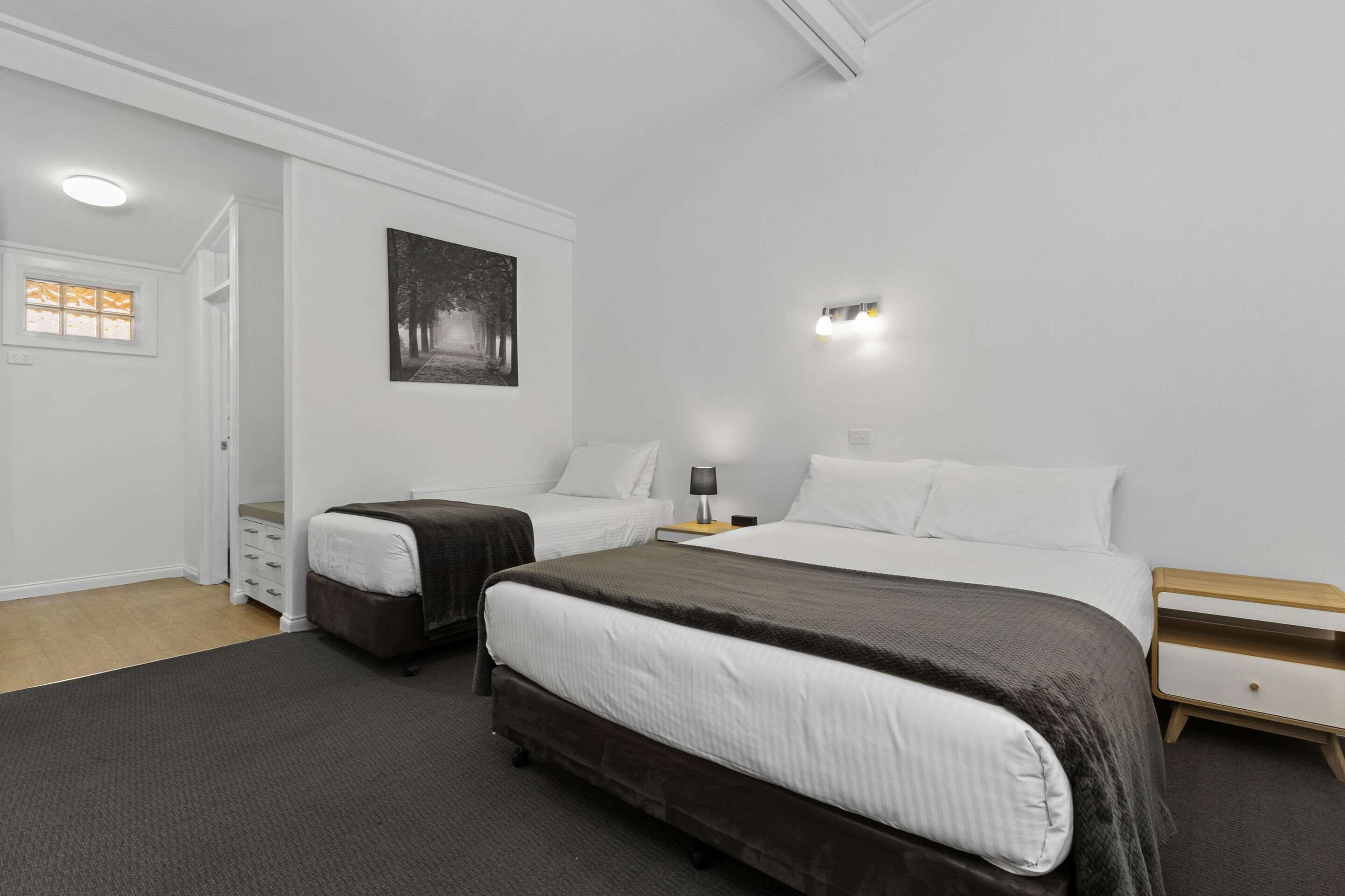 A hotel room with two beds, a bedside table with a lamp, a wall-mounted picture, and a door leading to a bathroom.