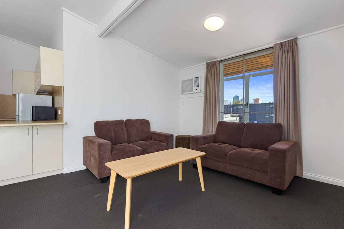 A modern living room with two brown sofas, a wooden coffee table, and a kitchen area with cabinets and a microwave.