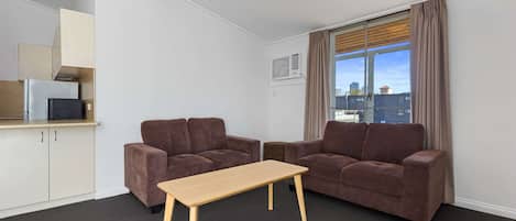 Una sala de estar moderna con dos sofás marrones, una mesa de centro de madera y una zona de cocina con armarios y un microondas.