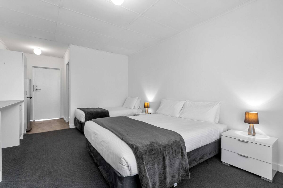 A modern hotel room with two single beds, a white wardrobe, a bedside table with a lamp, and a carpeted floor.