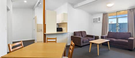 A modern living room with a wooden dining table, two sofas, and a small coffee table. There is a kitchenette in the background and a large window with curtains.