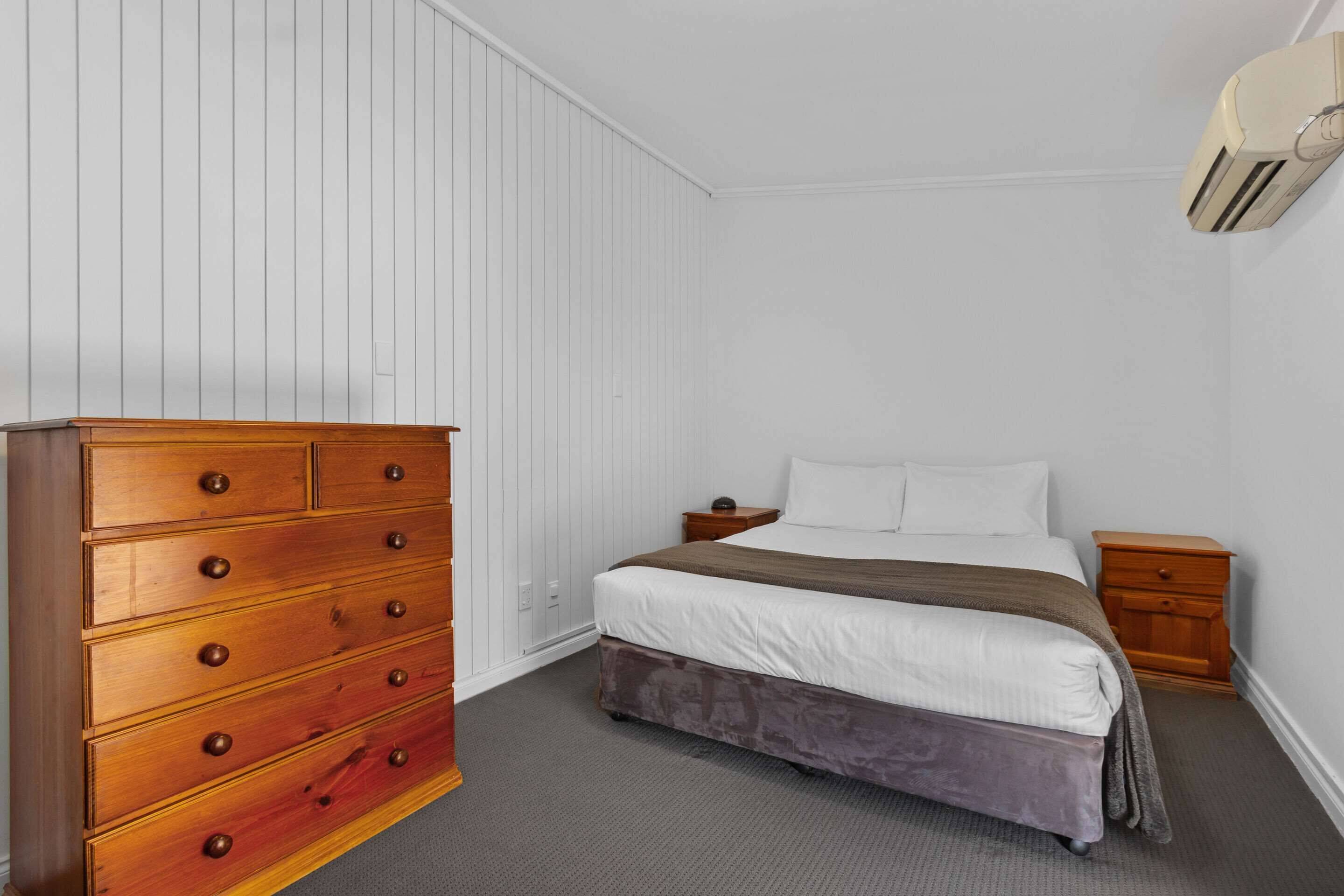 Une chambre à coucher avec une commode en bois, un lit avec une literie blanche, une table de chevet et un climatiseur fixé au mur.