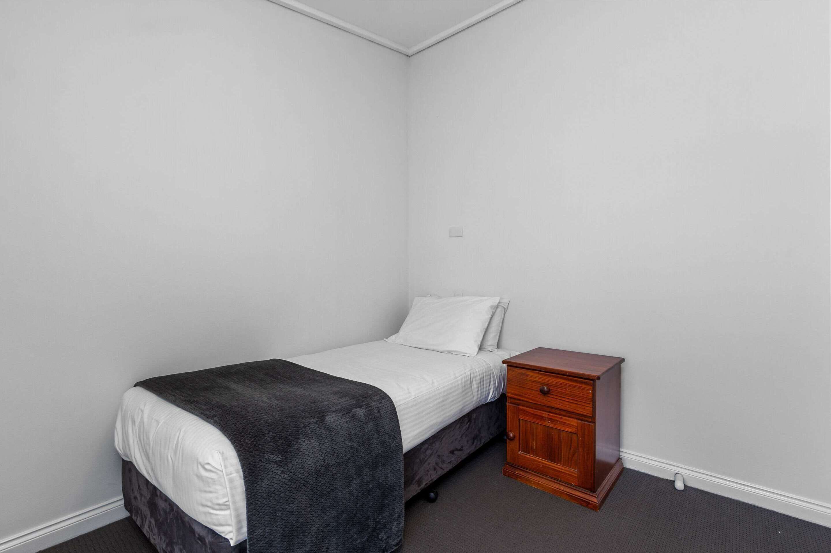A small, single-bed room with a dark-colored bedspread, a white pillow, and a wooden nightstand with drawers.