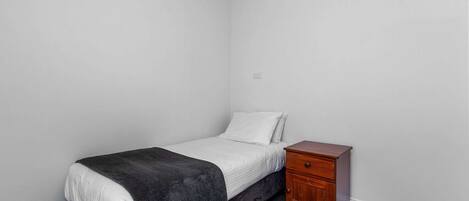 Una habitación pequeña con cama individual, colcha oscura, almohada blanca y mesita de noche de madera con cajones.