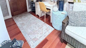 A room with a wooden door, a desk with a chair, a round mirror, a lamp, a patterned rug, and a sofa with cushions.