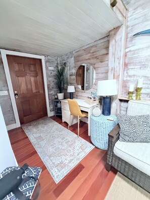 Une pièce avec une porte en bois, un bureau avec une chaise, un miroir rond, une lampe, un tapis à motifs et un canapé avec des coussins.