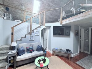 Un vaste espace de vie doté d’un haut plafond, d’un escalier, d’un canapé avec des coussins, d’une petite table et d’un téléviseur fixé au mur.